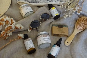 photo de plusieurs produits Comptoir des Huiles sur un drap en lin avec des fleurs séchées, des lunettes de soleil, un chapeau et une brosse à cheveux 